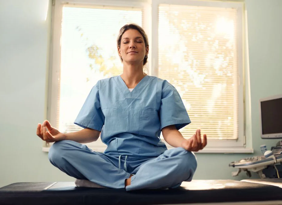 A nurse in scrubs sits cross-legged in a meditative position on a medical examination bed.