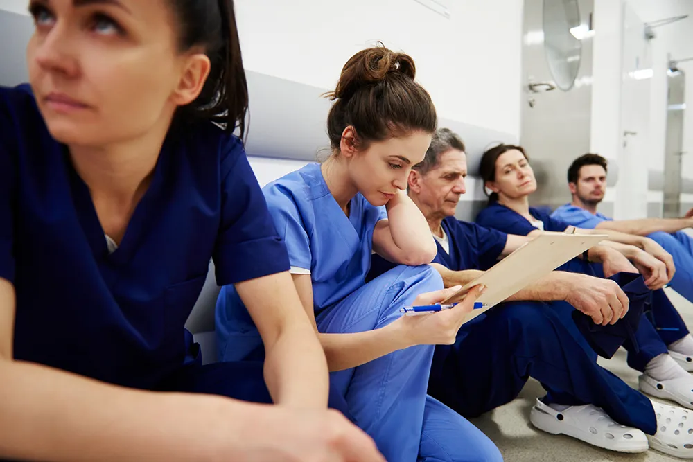 A group of nurses in scrubs seated together, lined up against a wall. One nurse in light blue scrubs is reviewing notes.