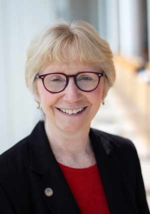 A headshot of Sally Thorne, smiling at the camera against a blurred backdrop. Sally is wearing glasses, has short blonde hair, and wearing a black blazer.