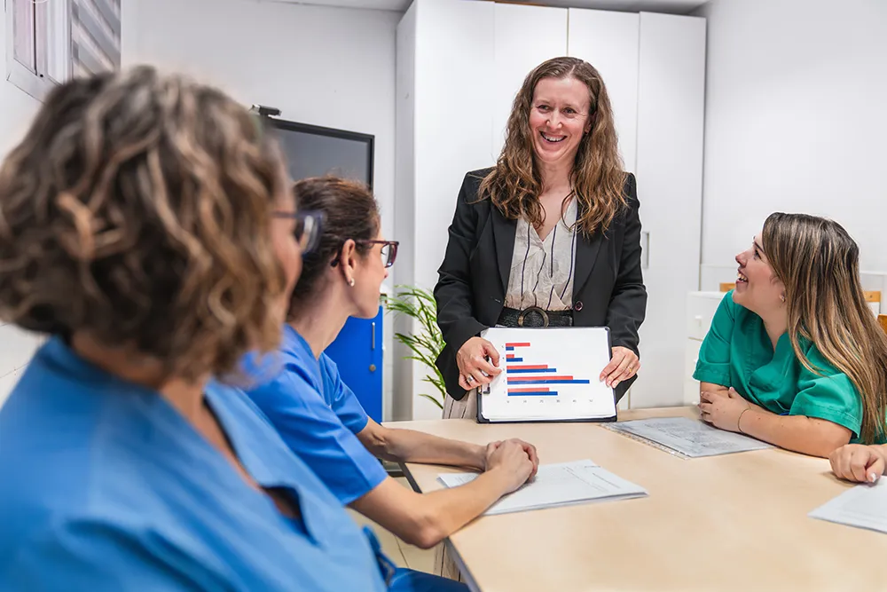 A health care presenter shows graphical data to a group of nurses during a leadership meeting in a boardroom.
