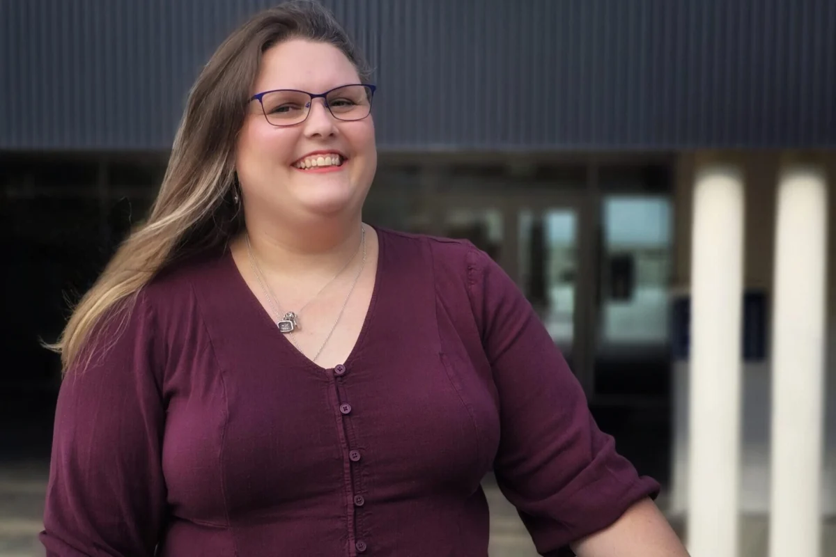 NNPBC-Westland Future Nurses Scholarship recipient, Linzi Irvine, stands outside a University of Victoria building. Linzi has long hair and is wearing a burgundy top and glasses.