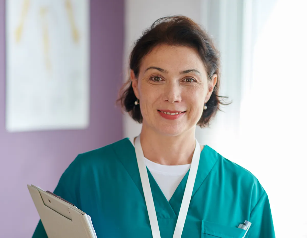 A headshot of a smiling nurse in scrubs, holding patient charts.