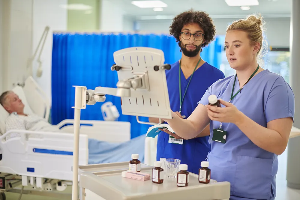Two nurses in scrubs stand near a patient's bedside, preparing and entering medication on the patient's medical chart, in an emergency room setting.