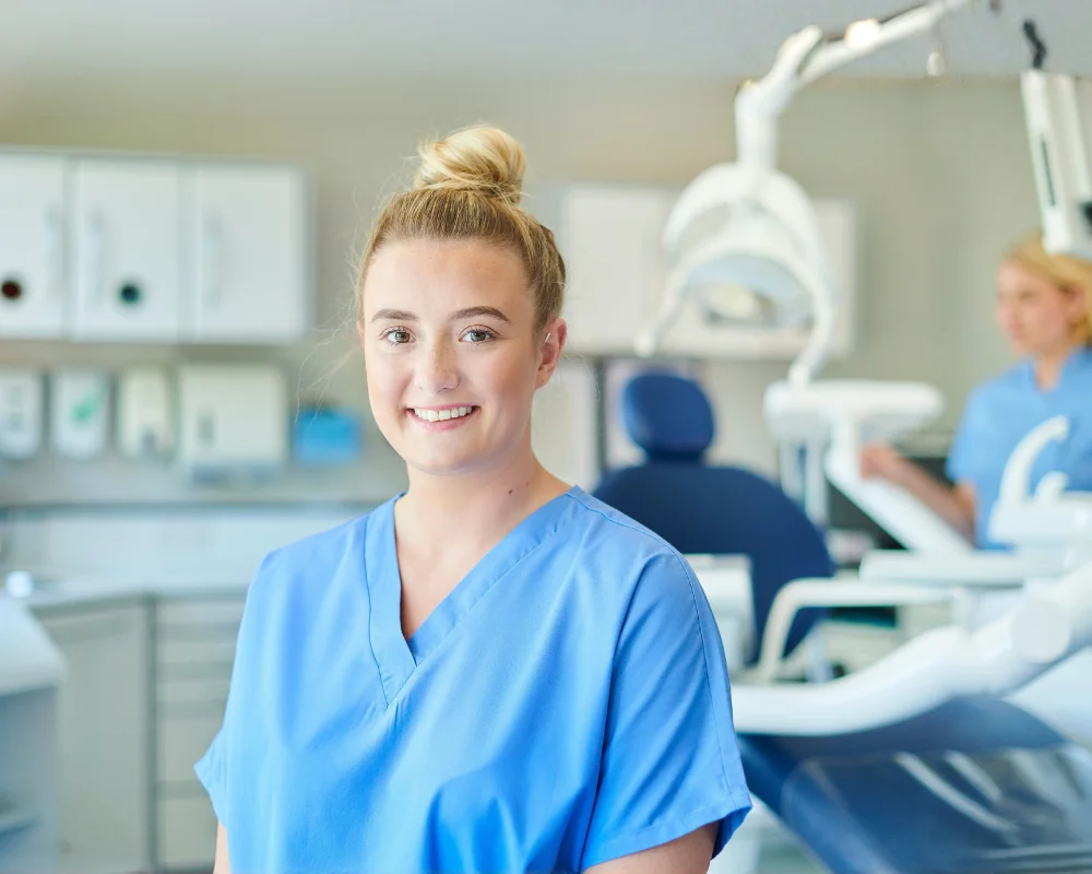 Young healthcare professional wearing blue scrubs, smiling in a modern dental clinic with dental equipment visible in the background.
