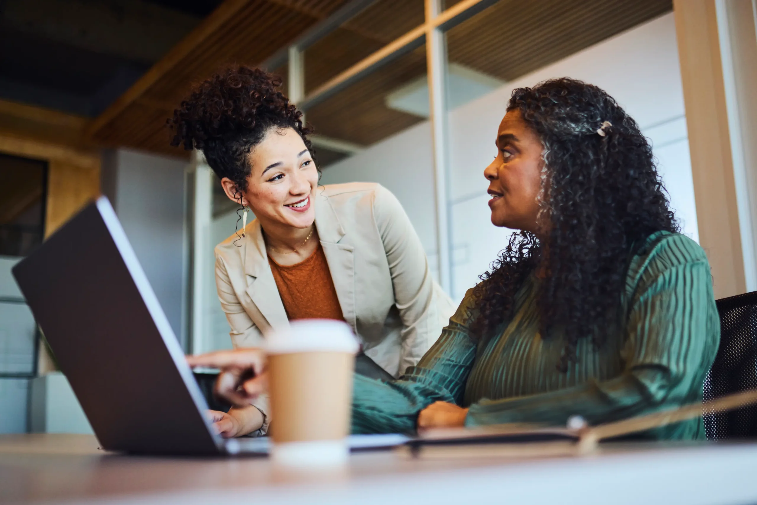 A young, smiling mixed-race businesswoman points at a laptop screen to a mature Black colleague during a collaborative discussion in a contemporary office setting.
